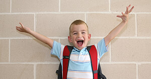 Young boy wearing a red backpack with arms raised and a huge smile on his face.