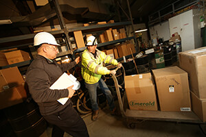 Two men wearing hard hats moving boxes in a warehouse.
