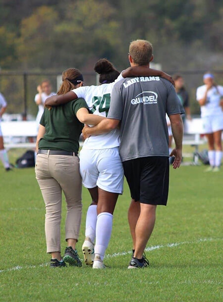 Jeena being led off the field after injury supported by her coach and an athletic trainer
