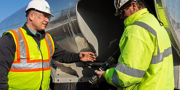 Two men wearing reflective gear and hard hats inspecting the fuel nozzle of an oil delivery truck.
