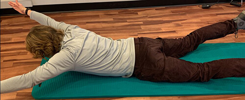 Man lying face down on an exercise mat, with arms outstretched and lifting his upper body.