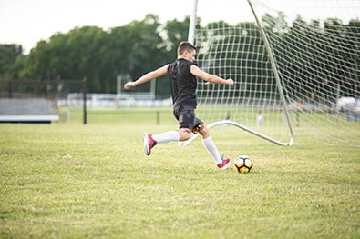 Young boy kicking soccer ball into net on field.