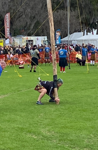 Cindy bends down to lift a caber in a green grass field in front of an audience.