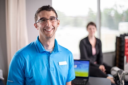 Male therapist wearing a teal blue shirt and name tag standing in front of a laptop on a desk..